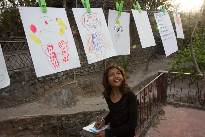 Drawings pegged on a line, a girl drawing below them, Rishikesh