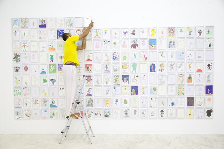 A man on a ladder hanging framed drawings on a white wall covered with aliens, Thaillywood