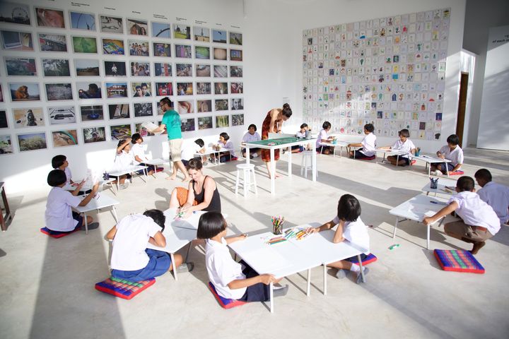 Children in school uniform drawing at low white tables in the gallery, the wall of aliens behind, Thaillywood